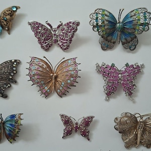 Può includere: Una collezione di undici spille a forma di farfalla in vari design, tra cui filigrana metallica, smalto colorato e accenti di strass scintillanti nelle tonalità blu, rosa, oro, argento e bronzo, disposte a griglia su una superficie piana.
