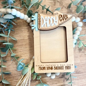 May include: Wooden photo frame with a banner that says "Best Daddy Ever" in white lettering on a light blue background. The frame has a tassel and says "Happy Father's Day 2024 from your biggest fan!"