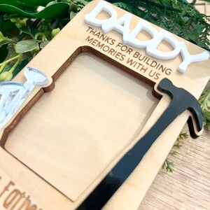 May include: Wooden photo frame with a hammer and nail cutout. The frame has the text "Daddy Thanks for building memories with us" and "Father's Day" at the bottom.