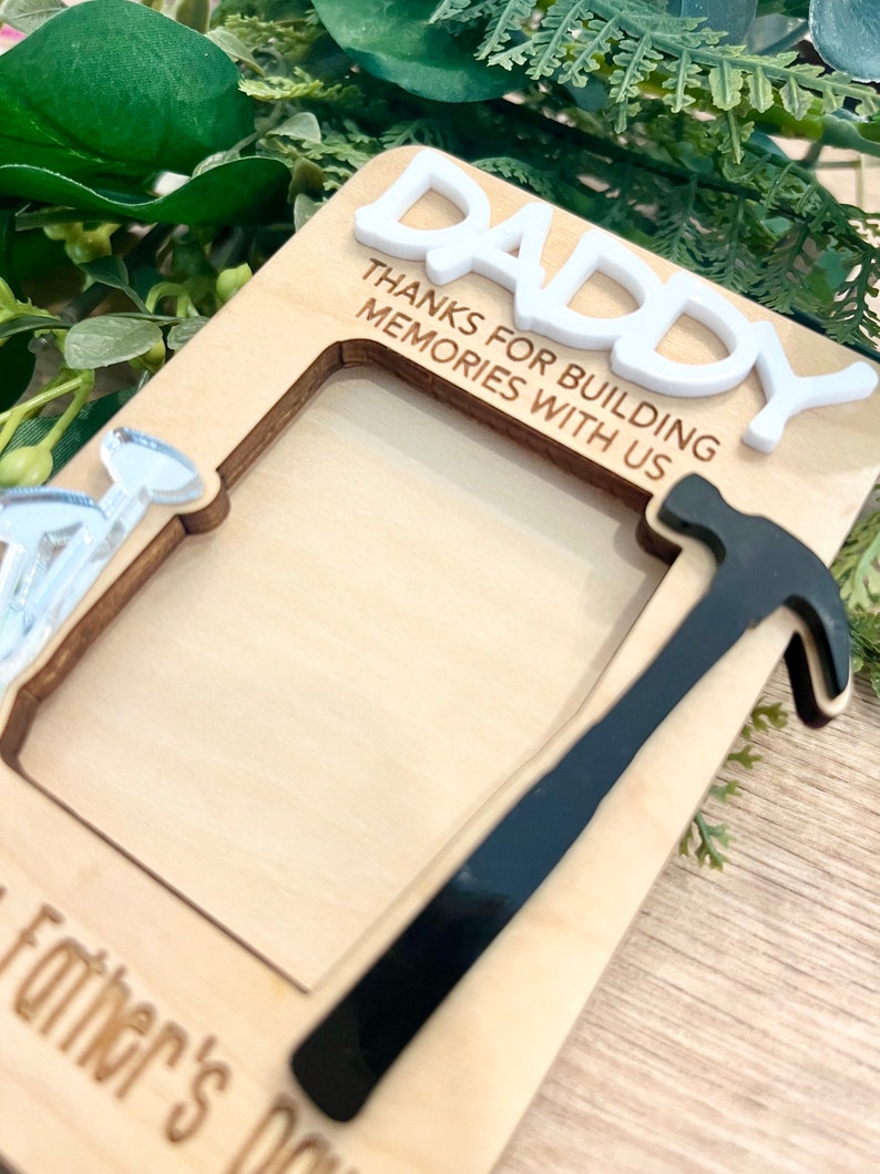 May include: Wooden photo frame with a hammer and nail cutout. The frame has the text "Daddy Thanks for building memories with us" and "Father's Day" at the bottom.