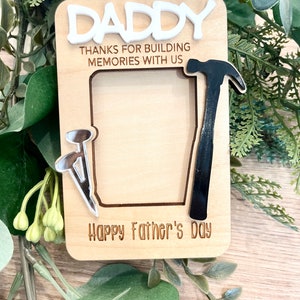 May include: A wooden photo frame with a cutout for a picture. The frame is decorated with a hammer, nails, and the text "Daddy Thanks for building memories with us Happy Father's Day".