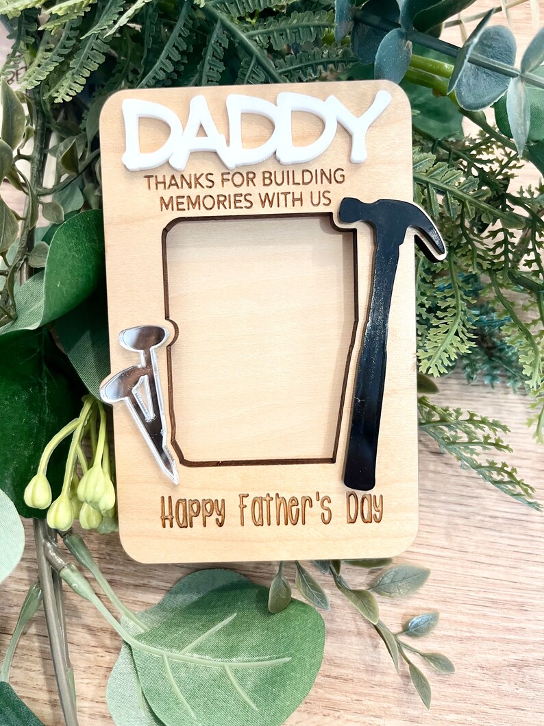 May include: A wooden photo frame with a cutout for a picture. The frame is decorated with a hammer, nails, and the text "Daddy Thanks for building memories with us Happy Father's Day".