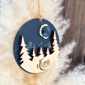 Peut inclure: Une décoration ronde en bois avec un fond noir et une lune jaune avec un cercle noir au centre. La décoration représente une silhouette de chaîne de montagnes et trois sapins. Le texte "ECLIPSE 04.08.2024" est imprimé au bas de la décoration.