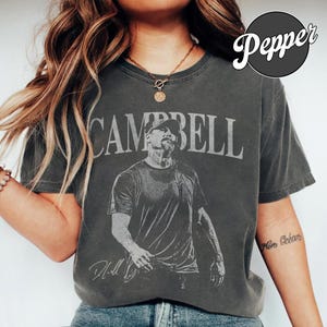 May include: A charcoal grey short-sleeve t-shirt featuring a graphic print of a man in a cap with the text "CAMPBELL" arched above and a signature below. The shirt has a crew neckline and a vintage-style washed finish.