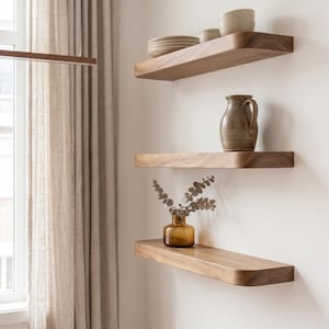 Op de afbeelding: Drie horizontale zwevende planken van natuurlijk hout met afgeronde randen, gemonteerd aan een witte muur. De bovenste plank bevat een stapel keramische borden en kommen. De middelste plank is voorzien van een bruine keramische kan. De onderste plank bevat een kleine amberkleurige glazen vaas met een takje gedroogde eucalyptus.
