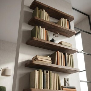 Könnte beinhalten: Vier dunkle, schwebende Holzregale an einer hellen Wand, jedes mit einer Sammlung von Büchern. Die Buchrücken sind beige, grün und orange. Eine kleine schwarze Vase und eine Skulptur sind ebenfalls vorhanden, was eine stilvolle Präsentation ergibt.