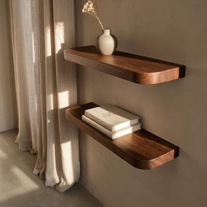 May include: Two dark brown wooden floating shelves with rounded edges mounted on a wall. The top shelf holds a small ceramic vase with a dried flower, and the bottom shelf holds two stacked books with light-colored covers and visible text.