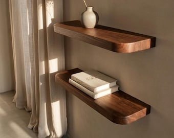 May include: Two floating wooden shelves with rounded edges are mounted on a neutral-toned wall. The top shelf holds a small, cream-colored vase with dried flowers. The bottom shelf displays two books. The shelves are made of dark wood.