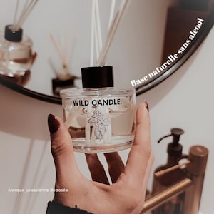 Peut inclure: Un diffuseur en verre transparent avec un bouchon noir et des bâtonnets blancs. La bouteille porte l'inscription "WILD CANDLE." et un motif de loup géométrique. Les mots "Base naturelle sans alcool" et "Marque jurassienne déposée" sont également visibles.