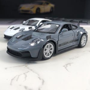 Pode incluir: Um carro modelo cinza com detalhes em preto e o texto "GT3 RS" na lateral. Há um carro branco atrás e um carro amarelo ao fundo. Os carros estão em uma superfície branca.