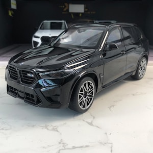 Op de afbeelding: Een glanzend zwart model van een BMW X5 M. De miniatuur-SUV heeft gedetailleerde kenmerken, waaronder een zwarte grille, koplampen en lichtmetalen velgen. Op de achtergrond is een andere witte auto zichtbaar. Het model wordt gepresenteerd op een marmeren oppervlak.