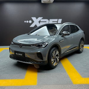 Puede incluir: Un modelo de coche gris Volkswagen ID.4 Crozz con techo negro, exhibido sobre una superficie gris con marcas amarillas. El coche tiene faros detallados, una parrilla delantera y el texto "ID.4 CROZZ" en la parte delantera.