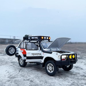 Könnte beinhalten: Weißes und schwarzes Toyota Land Cruiser Modell mit geöffneter Motorhaube, Ersatzreifen und Dachgepäckträger. Das Modell hat schwarze Reifen, einen roten Streifen und das Wort "TOYOTA" an der Seite. Es hat eine Lichtleiste auf dem Dach und gelbe Lichter vorne.