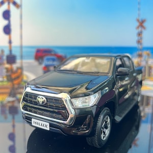 May include: A black Toyota Hilux scale model pickup truck featuring a chrome-accented front grille, clear headlights, and a license plate with the text HILLUX. The vehicle is positioned on a reflective surface with a blurred coastal background.