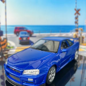 Könnte beinhalten: Ein blaues Modellauto aus Druckguss mit einer schlanken Coupé-Karosserie, klaren Scheinwerfern, einem Frontgrill mit kleinem Emblem und silbernen Mehrspeichenfelgen. Das Fahrzeug steht auf einer reflektierenden Oberfläche und zeigt sein Design.