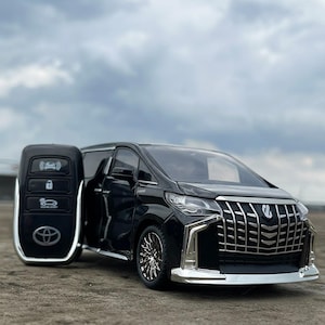 Puede incluir: Un modelo negro de Toyota Alphard con un llavero a juego. La furgoneta tiene detalles cromados en la parrilla delantera y el parachoques. El llavero es negro con detalles plateados y el logotipo de Toyota.