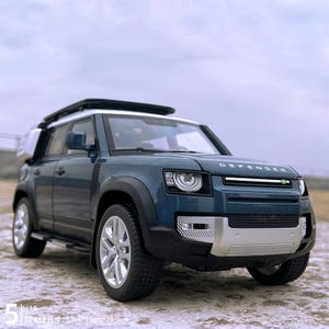 Puede incluir: Maqueta de un Land Rover Defender de color verde azulado con techo blanco y detalles negros. El modelo tiene ruedas plateadas y una parrilla delantera detallada. El texto "DEFENDER" es visible. Escala 1:18, dimensiones 32cm x 13cm x 11.5cm.