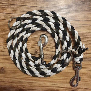 Puede incluir: Una correa para perro trenzada en negro, blanco y verde con un clip plateado y un anillo plateado.