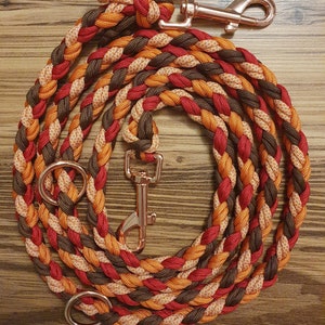 Puede incluir: Una correa de perro trenzada en naranja, rojo y marrón con un cierre de metal dorado rosa y dos anillos de metal dorado rosa.