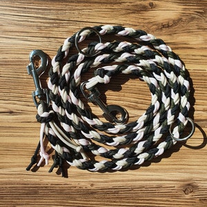 Puede incluir: Una correa para perro trenzada en negro, blanco y verde con dos clips plateados y un anillo plateado.
