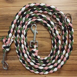 Puede incluir: Una correa para perro trenzada con cuerda negra, blanca, rosa y verde. La correa tiene dos clips metálicos y dos anillos metálicos.