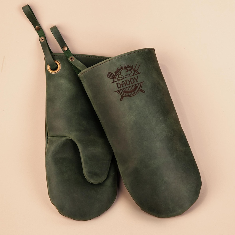 May include: A pair of green leather oven mitts with a hanging loop. The right mitt has the text "Daddy" engraved on it with a chef hat and crossed grilling utensils.