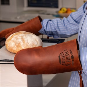 May include: A pair of brown leather oven mitts with the text "Cherie's Kitchen" and a rolling pin design. The mitts are on a baking sheet with a loaf of bread.
