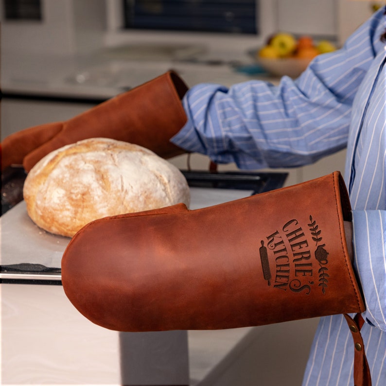 May include: A pair of brown leather oven mitts with the text "Cherie's Kitchen" and a rolling pin design. The mitts are on a baking sheet with a loaf of bread.