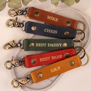 May include: A selection of leather keyrings in shades of brown, navy, green, red, and orange. Each keyring has a gold-coloured clasp and gold-embossed text, including "MIKE", "CHRIS", "BEST DADDY", "BEST MOM", and "LILY".