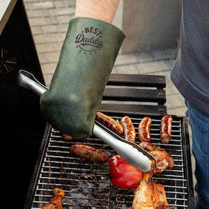 May include: A green leather grilling mitt with the text "BEST Daddy" in a brown font. The mitt is being used to hold a pair of tongs to flip food on a grill.