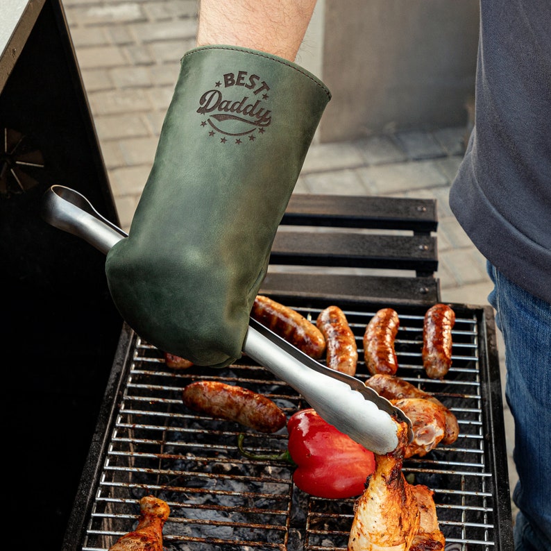May include: A green leather grilling mitt with the text "BEST Daddy" in a brown font. The mitt is being used to hold a pair of tongs to flip food on a grill.