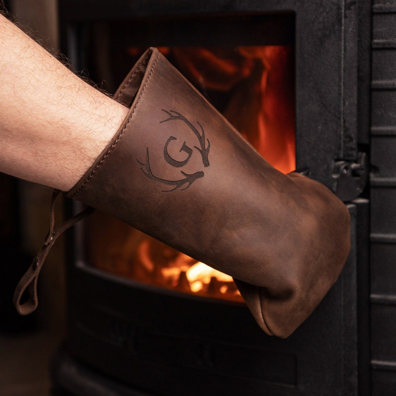May include: A brown leather oven mitt with a deer antler design and the letter "G" in the center. The mitt is being used to open a wood-burning stove.