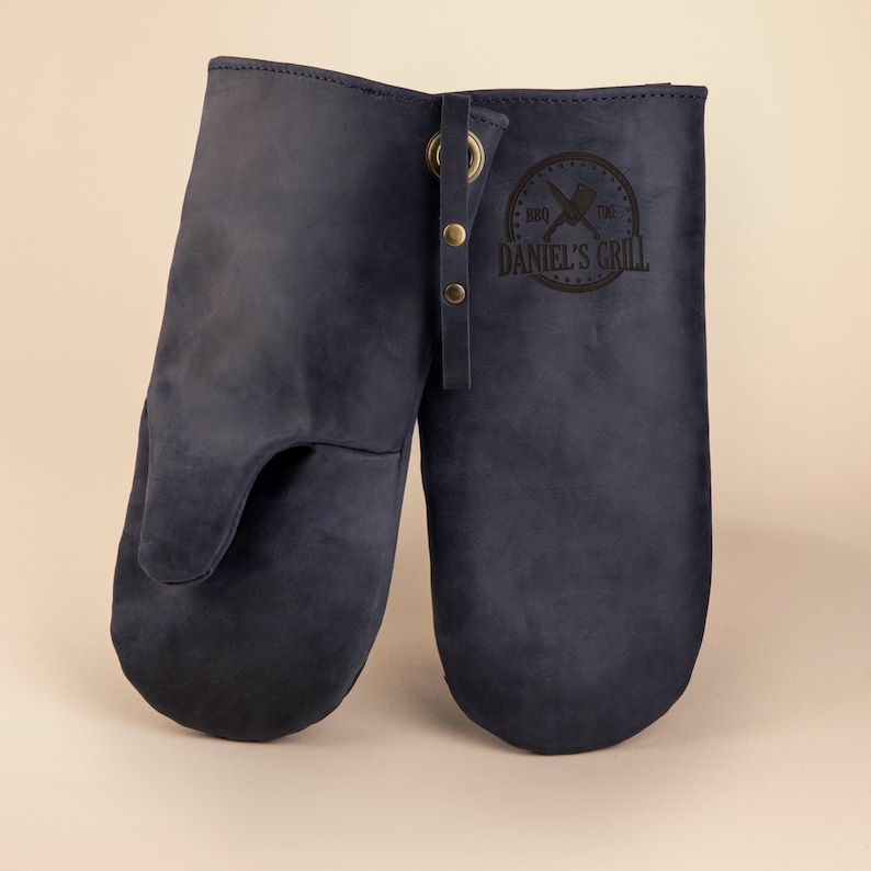 May include: A pair of blue leather oven mitts with a black leather patch that says "BBQ Time Daniel's Grill".