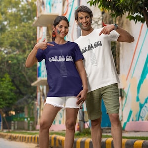 Puede incluir: Dos personas vistiendo camisetas con un diseño gráfico de un edificio con el texto "Capital Cell" y las palabras "We built it different" debajo. La persona de la izquierda lleva una camiseta azul marino y la persona de la derecha lleva una camiseta blanca. Ambos están de pie en una calle con una acera rayada amarilla y negra.