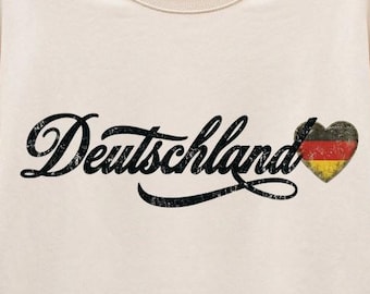 May include: A cream-colored crewneck sweatshirt featuring the word Deutschland in a black, distressed, cursive font. To the right of the text is a small heart graphic filled with the black, red, and gold horizontal stripes of the German flag.