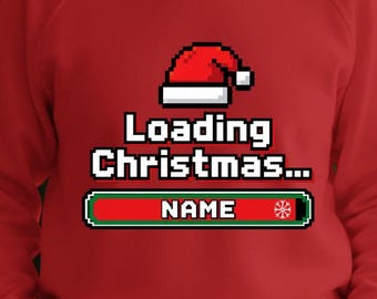 May include: A red long-sleeve sweatshirt featuring a pixelated graphic of a Santa hat above the text Loading Christmas and a progress bar with the word NAME and a snowflake icon. The design is centered on the front of the garment.