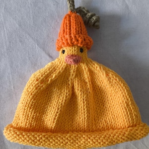 May include: A yellow knitted beanie featuring a duck-shaped top with a small orange beak, two black eyes, and a ribbed orange hat worn by the duck. The main body of the beanie is constructed with a vertical ribbed knit pattern and a rolled brim.