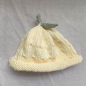 May include: A knitted baby hat in a pale yellow color, resembling a lemon. The hat features a small, light green leaf detail at the top. The hat is made of soft yarn and has a ribbed brim.