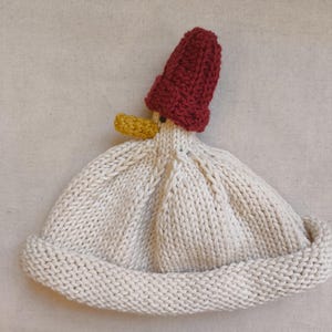 May include: A knitted hat in the shape of a duck. The hat is primarily off-white with a red cap and a yellow beak. The hat is made of thick yarn and has a folded brim.