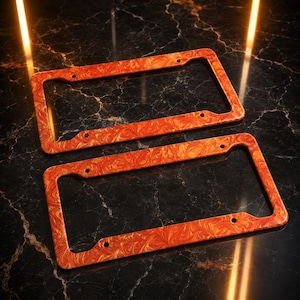 May include: Two rectangular license plate frames featuring a vibrant orange and red marbled pattern. Each frame has four pre-drilled mounting holes positioned at the corners, designed to secure a standard license plate to a vehicle exterior.