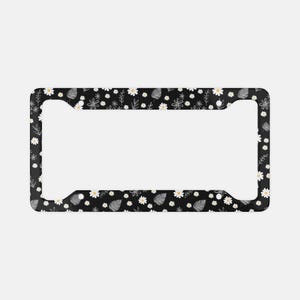 May include: A rectangular license plate frame featuring a black background with a repeating pattern of white daisies with yellow centers, interspersed with small black and white line-drawn leaves and botanical sprigs across the entire surface.