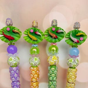 May include: Four decorative beaded items with a koi pond theme. Each has a jeweled top, a green lily pad with a small koi fish, and a colorful beaded stem. The stems feature various colors, including purple, yellow, and green, with floral and shell-like details.