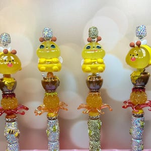 May include: Four decorative pens with bee-themed designs. Each pen features a yellow bee figurine, a honey pot, and a sparkling crystal top. The pen barrels are embellished with rhinestones and additional bee accents.