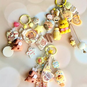 May include: Three decorative keychains featuring yellow rings and various charms including capybaras, Winnie the Pooh, and Hello Kitty. Charms include hearts, stars, and food items with text such as Capybara, play cute, and honey.