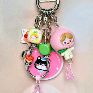 May include: A silver-toned metal keychain featuring a pink circular charm with a black cat illustration and the word HAPPY, a pink peach-shaped character charm, a yellow cube bead with a cat face, a green cube bead, and a translucent star charm.