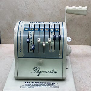 May include: Vintage Paymaster cash register in a light green and white color scheme. The register features dollar and cent indicators, a handle, and the word "Paymaster" in cursive. The register has a warning label at the bottom.