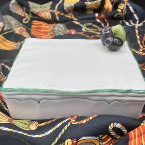 May include: A white ceramic box with a green trim and a small olive design on the top. The box is sitting on a black and gold patterned fabric.