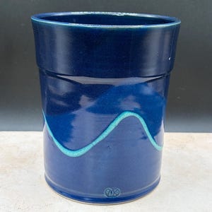 May include: A cylindrical ceramic tumbler with a glossy deep blue glaze. The vessel features a single, raised, wavy line in a contrasting light turquoise color that wraps around the middle. A small, circular maker's mark is embossed near the base.