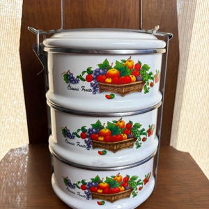 May include: A three-tiered white metal food carrier with a silver handle and locking mechanism. Each of the three cylindrical containers features a colorful graphic of assorted fruits in a woven basket with the text Classic Fruits printed below.