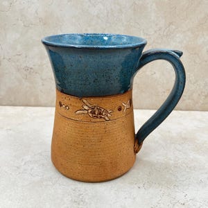 May include: A handmade ceramic mug with a blue interior and handle, and a tan exterior. The mug features a decorative band with a turtle and star design. The mug is approximately 5 inches tall.